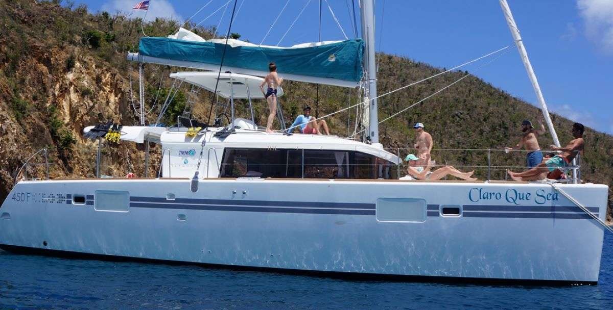 Sailing catamaran named 'Claro Que Sea' anchored near rocky coast with several people sunbathing and lounging on deck.