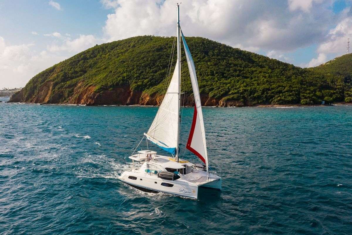 White catamaran sailing in blue sea past a green, rocky island under a partly cloudy sky.