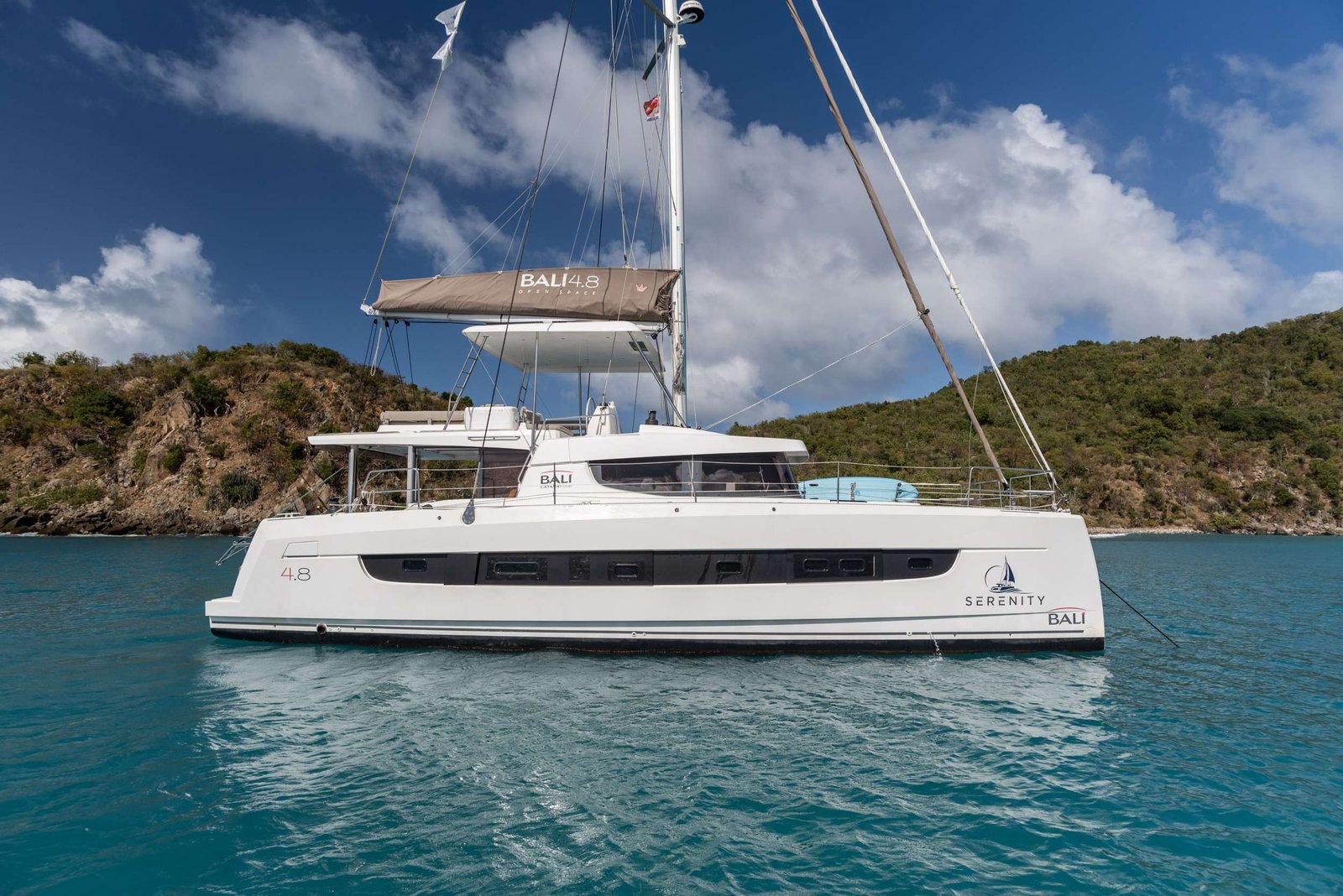 Catamaran yacht named Serenity floating in clear turquoise water with rocky coastline and blue sky in the background. The hull reads Bali 4.8 and Bali logo is visible.