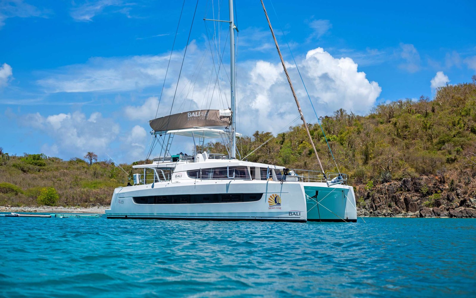 White catamaran sailboat anchored near a rocky tropical shore with blue sky and scattered clouds in the background