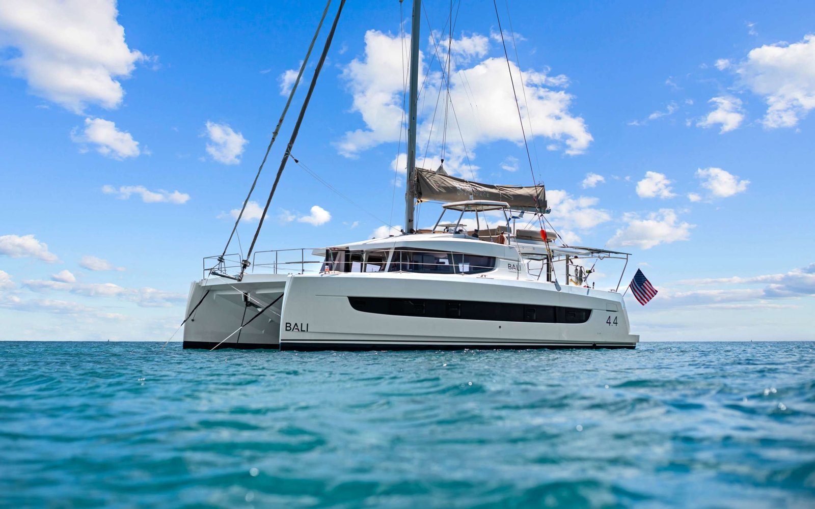White catamaran yacht anchored on calm turquoise water with a bright blue sky and scattered clouds; American flag at the stern.