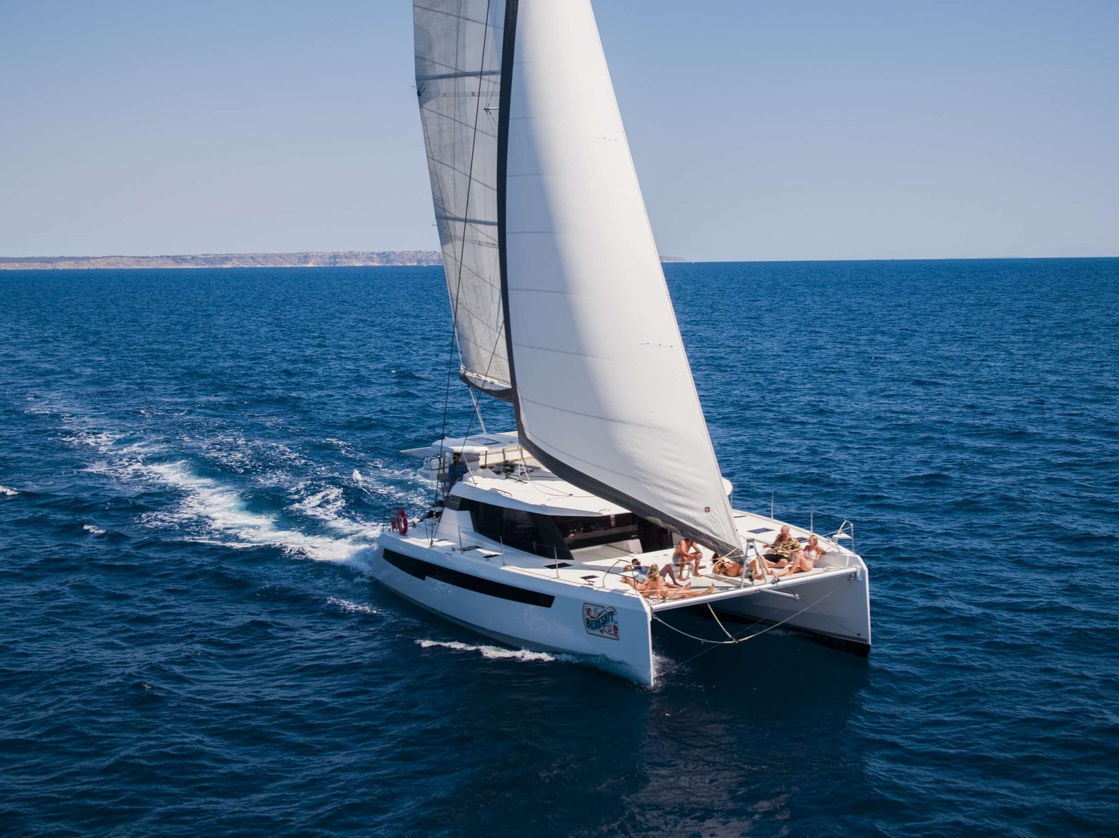 Sailing yacht with white sails on blue sea; several people on the deck enjoying the ride.