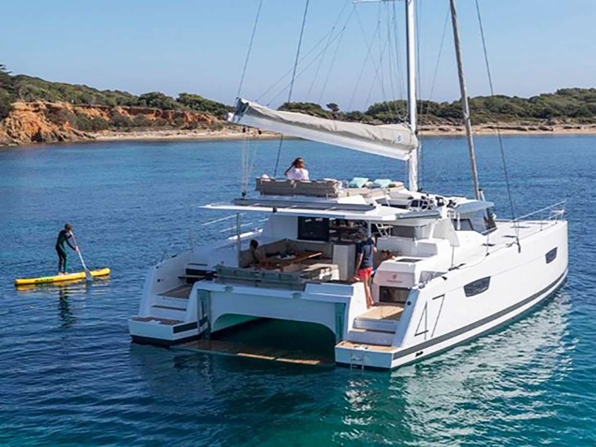 White catamaran anchored in turquoise water near a rocky shoreline, with a person paddling a yellow stand-up paddleboard nearby.