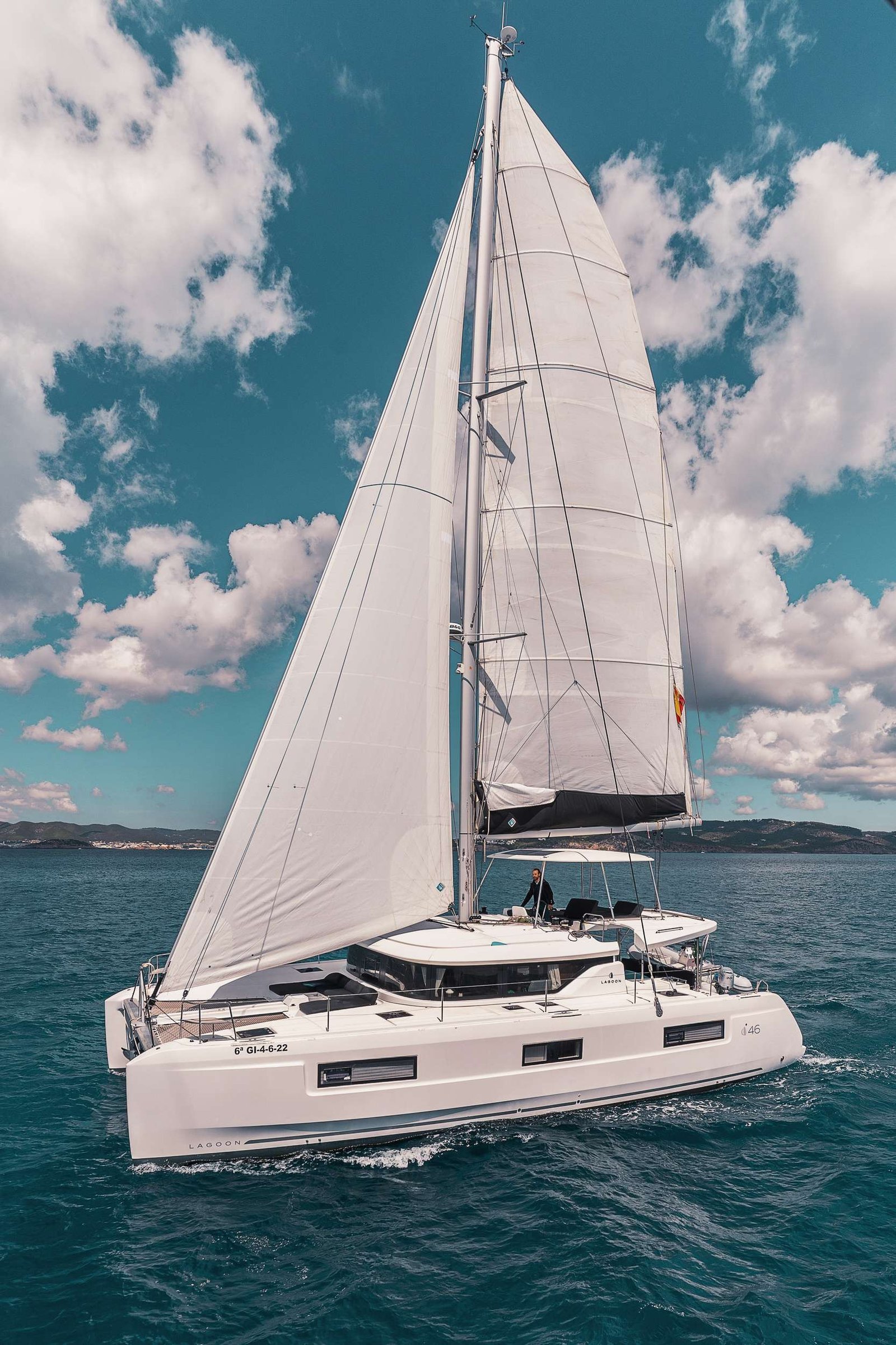 White Lagoon catamaran sailing on turquoise water with raised sails and a blue sky, a person on deck at the stern
