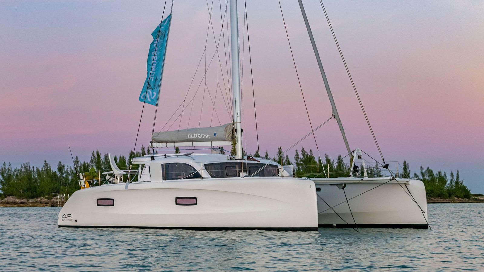 Catamaran sailboat on calm water at sunset with pastel pink and blue sky and treeline in the distance.