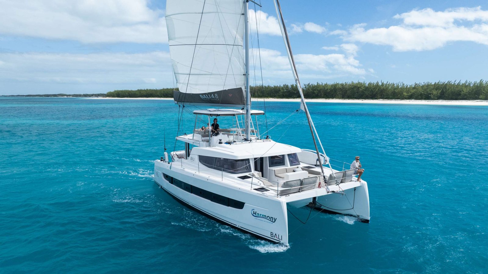 White catamaran sailing boat with two hulls on clear turquoise water near a sandy island and green trees; two people relaxing on deck under a large sail.