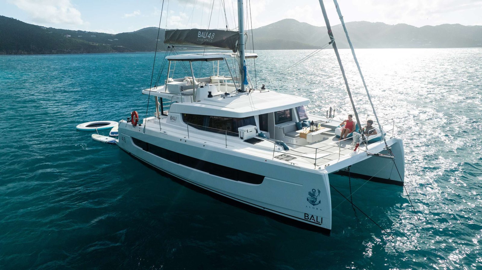 White catamaran yacht anchored in calm turquoise water, two people lounging on the stern as hills rise in the distance.