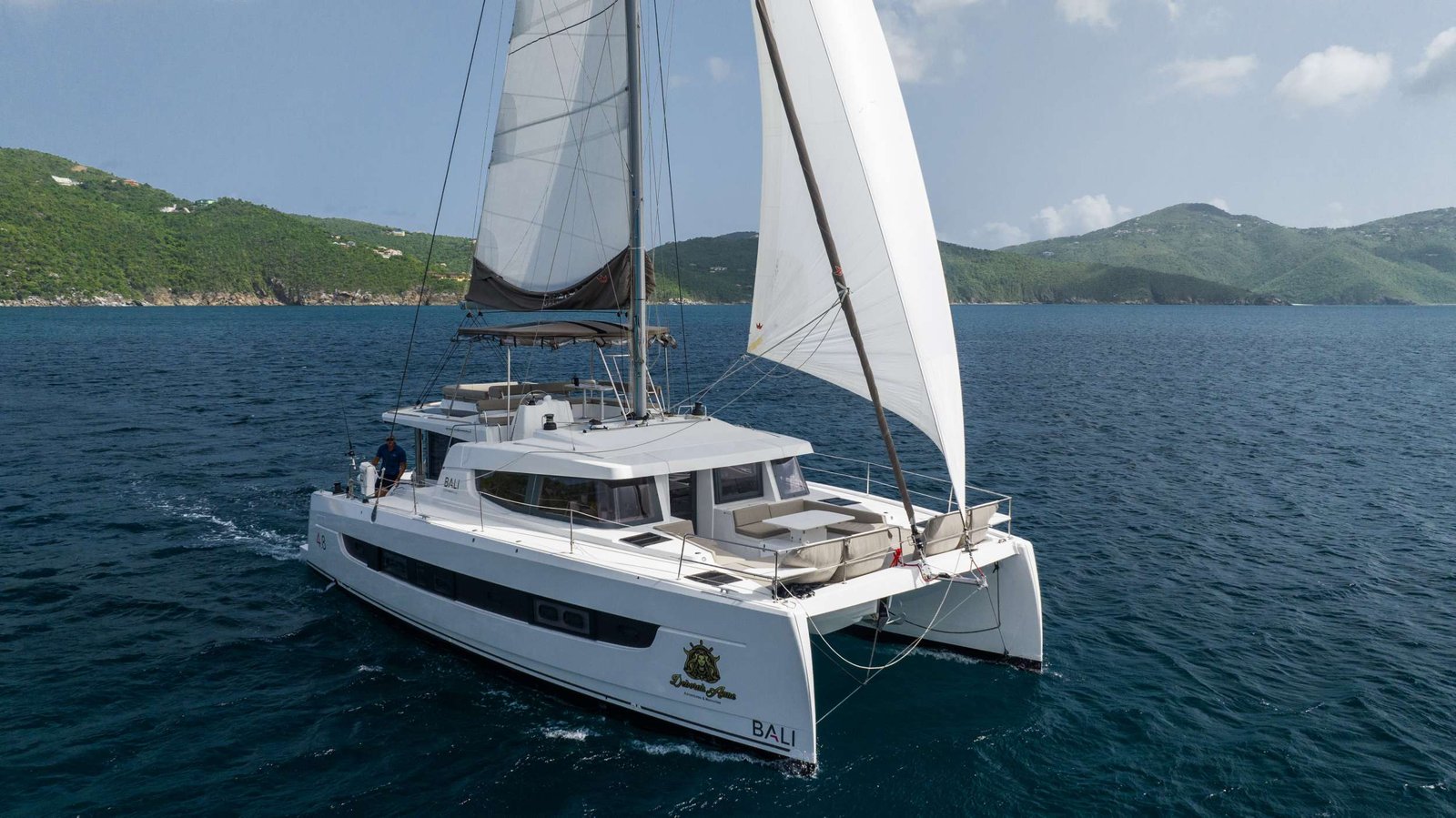 White catamaran sailing in clear blue sea with green hills in the background; two people on deck.