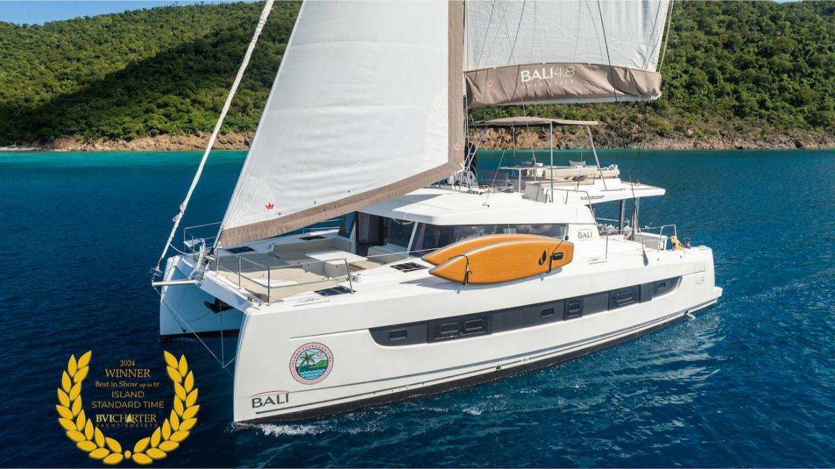 White catamaran Bali sailing yacht on blue water, with an orange deck board and a forested coastline in the background, award emblem at lower left.
