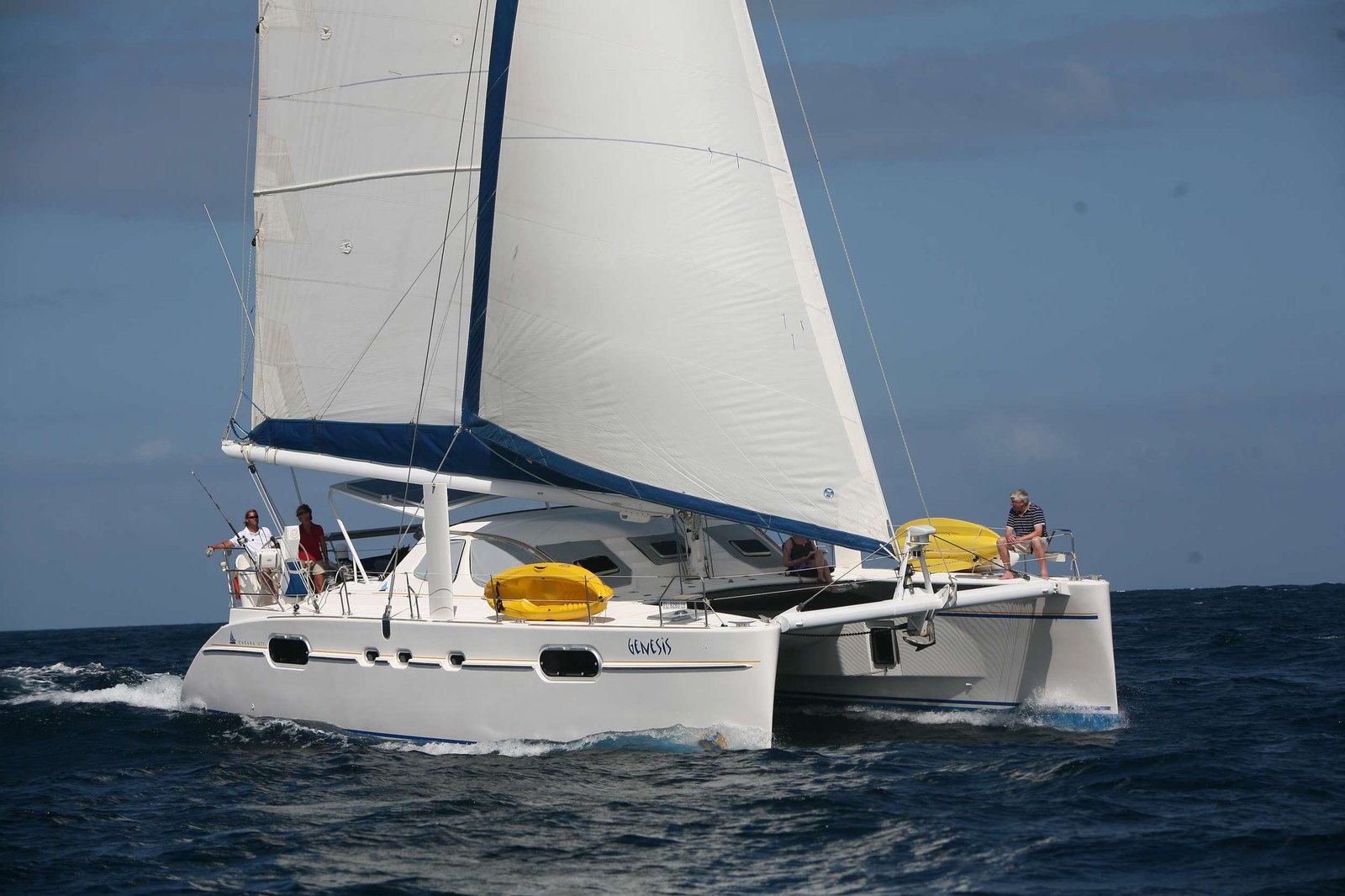 Catamaran sailboat named Genesis cutting through the sea with people on deck under white sails and blue sky.