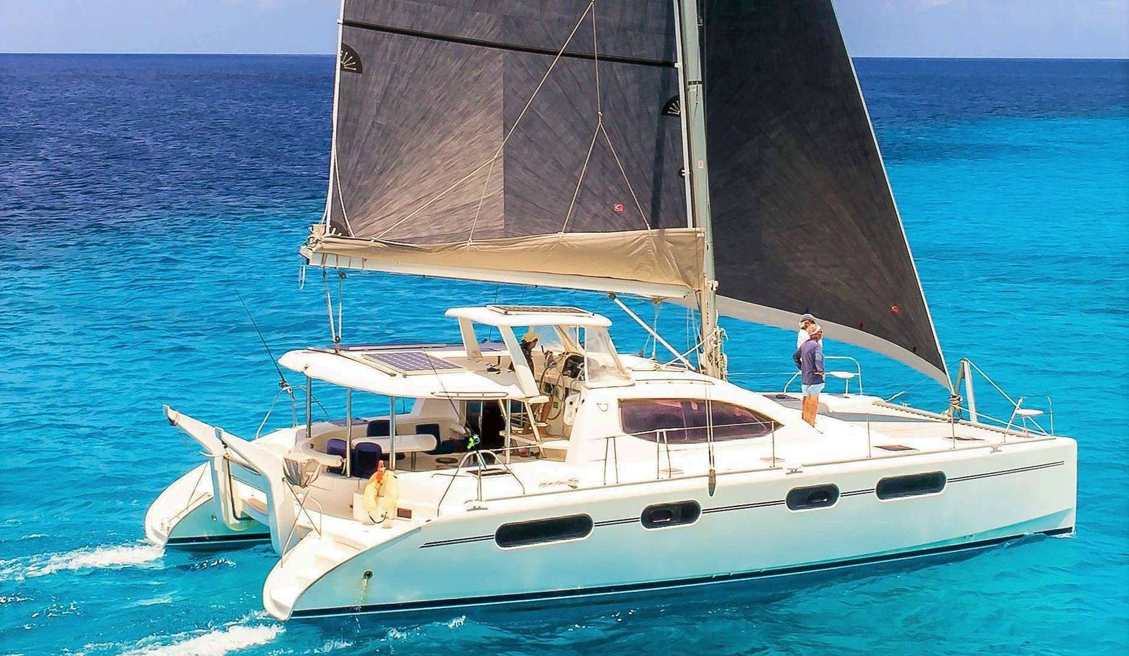 White catamaran sailing yacht with a large dark sail, a person standing on the deck against vibrant turquoise water.
