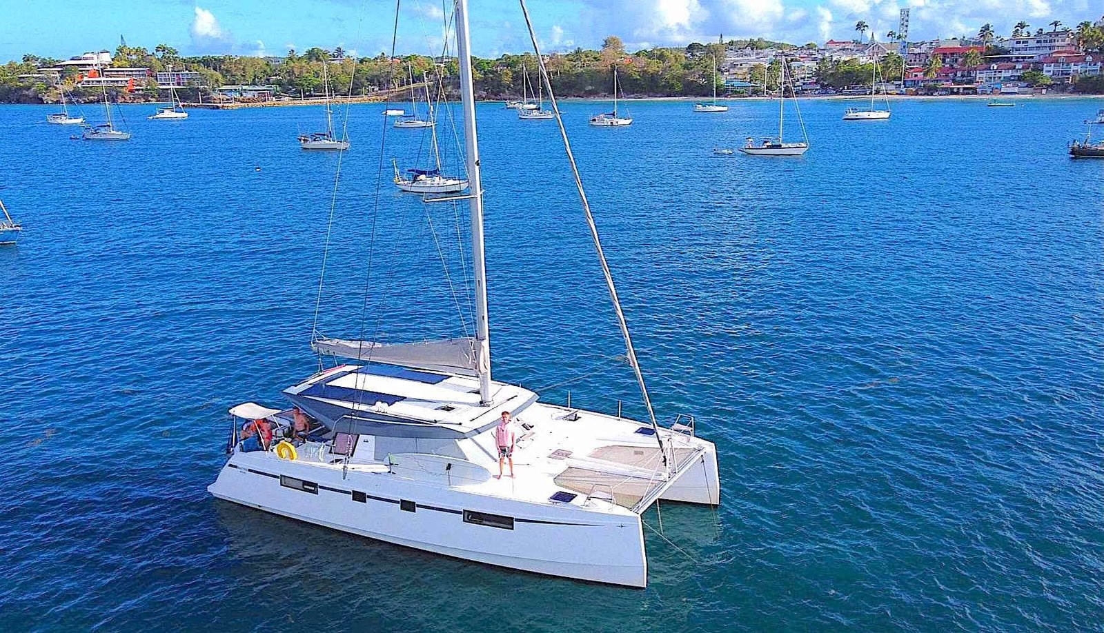 White catamaran sailboat with a person on deck in bright blue water, anchored near a shoreline with houses and other boats nearby