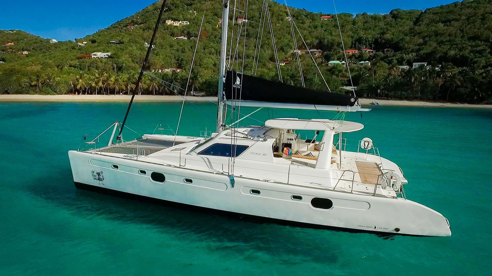 White catamaran anchored in turquoise water near a tropical beach with palm trees and green hills in the background.