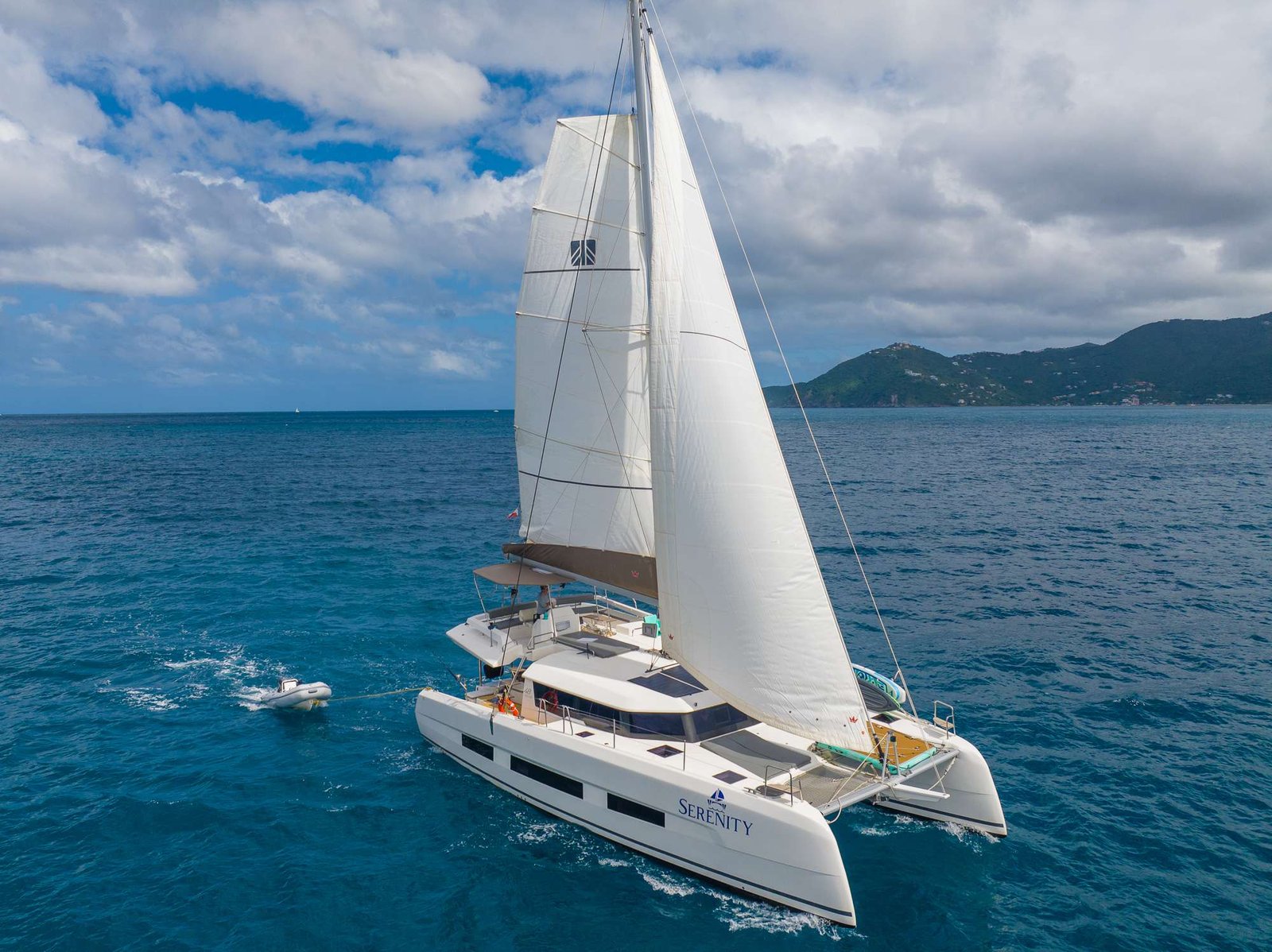 White sailing catamaran on turquoise sea with large sails and distant green hills under a partly cloudy sky.