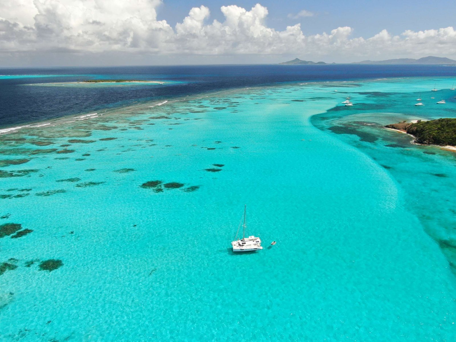 Aerial view of a turquoise lagoon with anchored sailboats and distant islands on the horizon under a cloudy sky