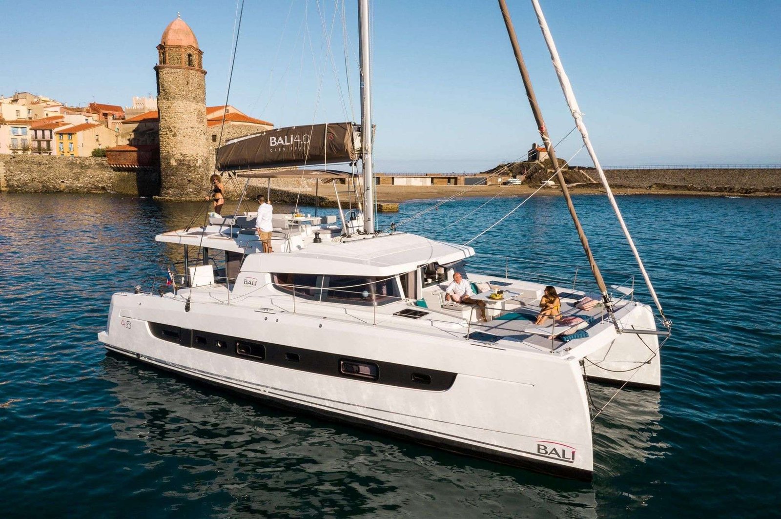 White Bali catamaran anchored near a stone harbor wall with people relaxing on deck.