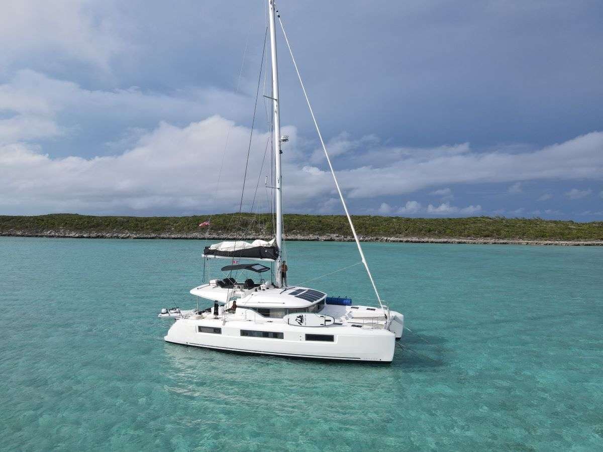 White catamaran sailboat anchored in clear turquoise water with a distant rocky shoreline under a partly cloudy sky