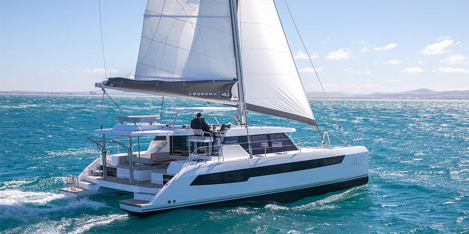 Modern white catamaran sailing on blue ocean with large white sails and a crew member at the helm; coastline in the distance under a clear sky