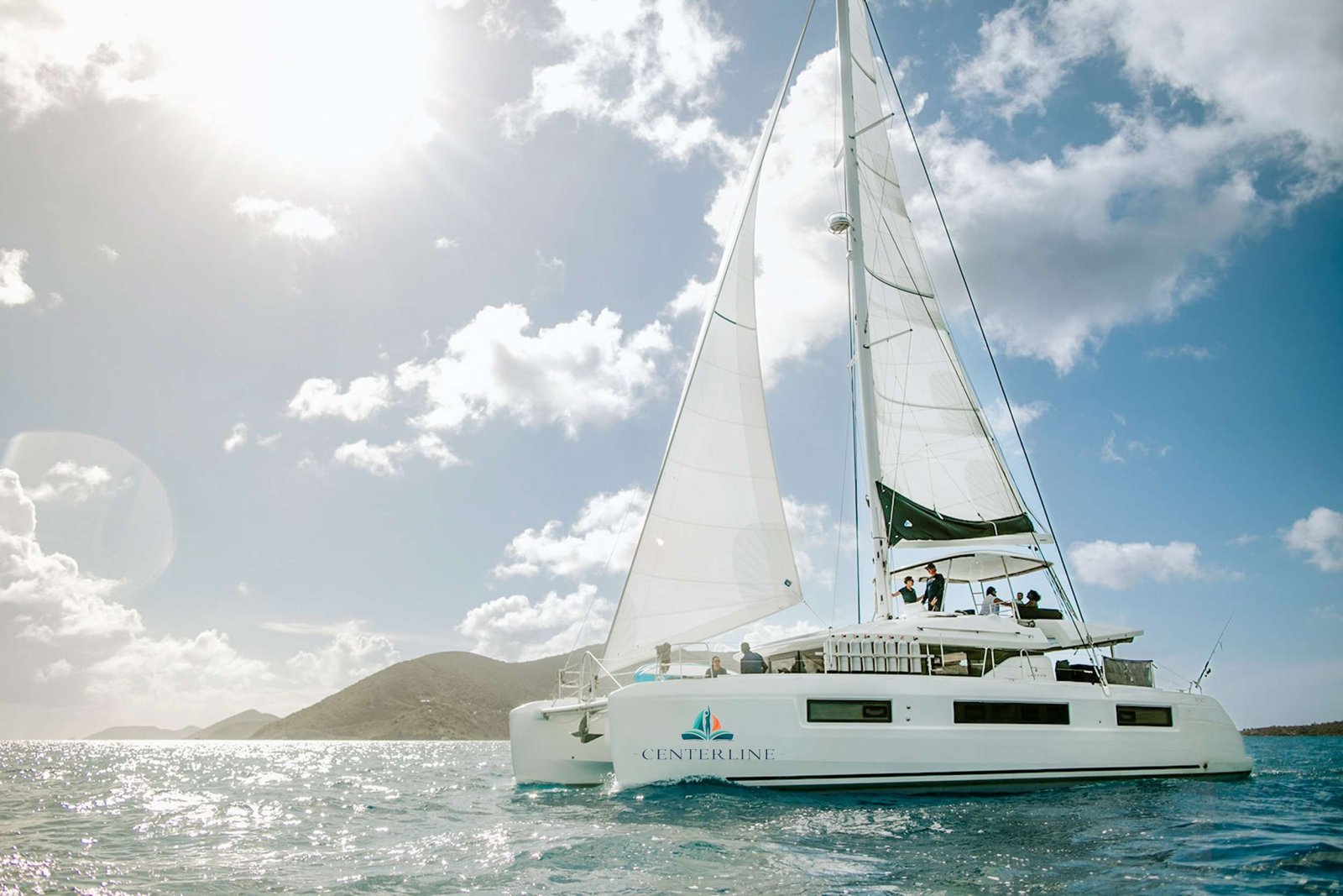 Sailboat with white sails and passengers on the deck gliding over turquoise water near a rocky coastline under a bright sky; Centerline logo on hull.