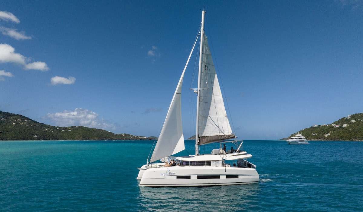 White catamaran sailing on clear turquoise sea with hills in the distance and a distant yacht visible on the right side.