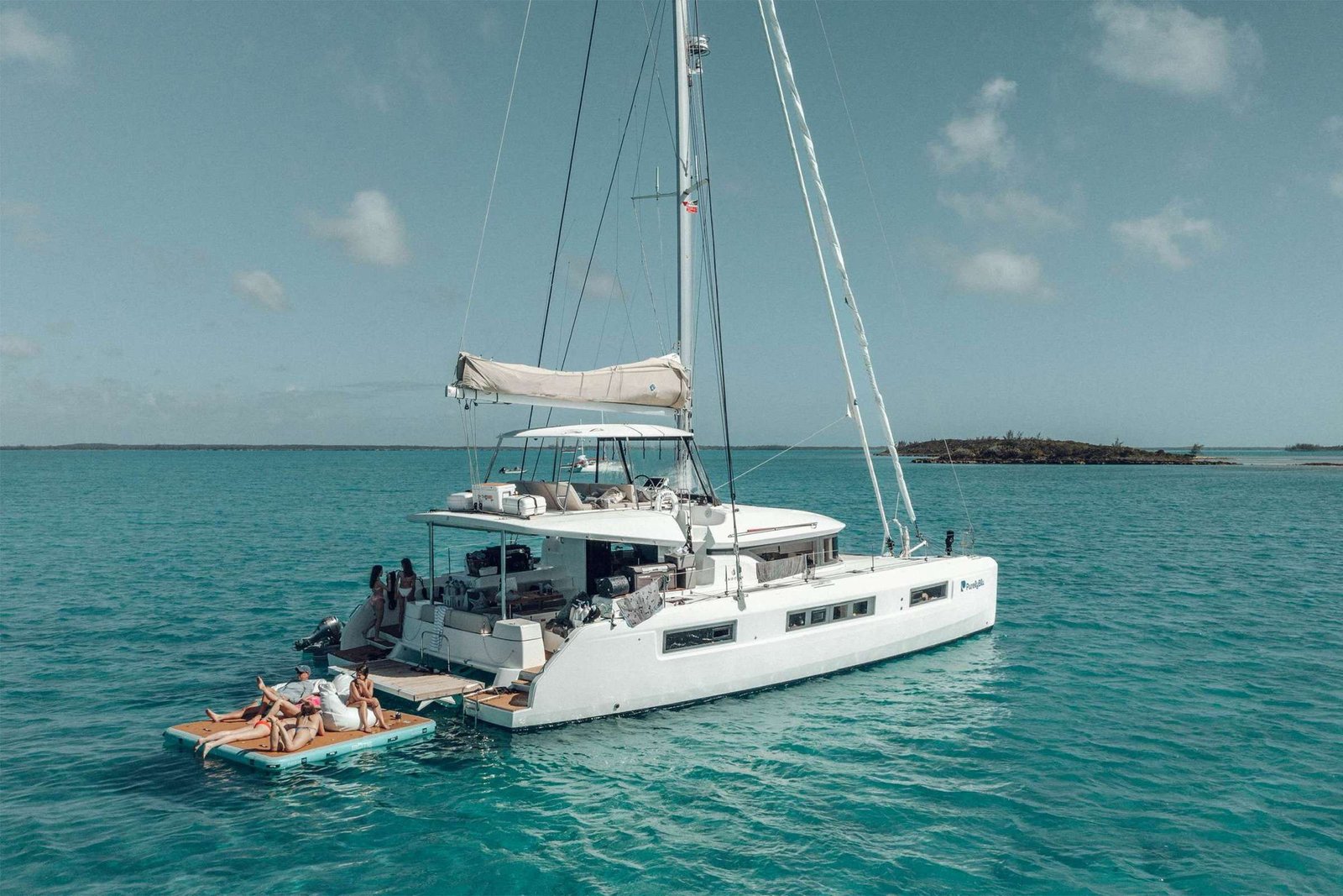 White catamaran anchored in turquoise water with a floating orange platform where people sunbathe nearby. The open deck shows seating and gear under a blue sky.