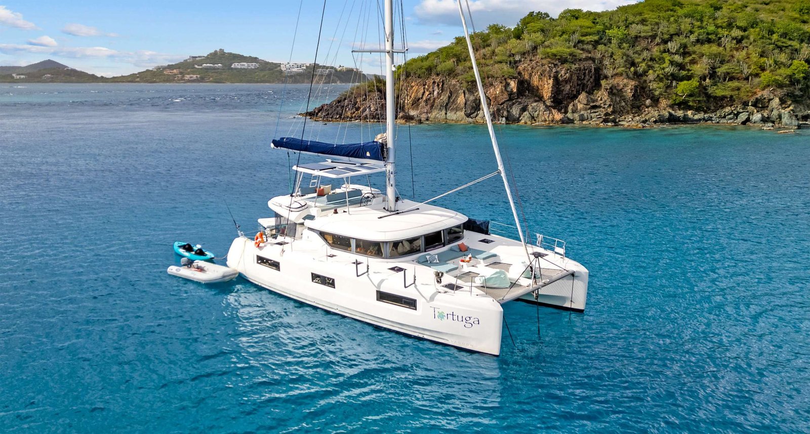 White catamaran named Tortuga anchored in a turquoise bay beside a rocky, tree-covered coastline; small kayaks attached to the side.