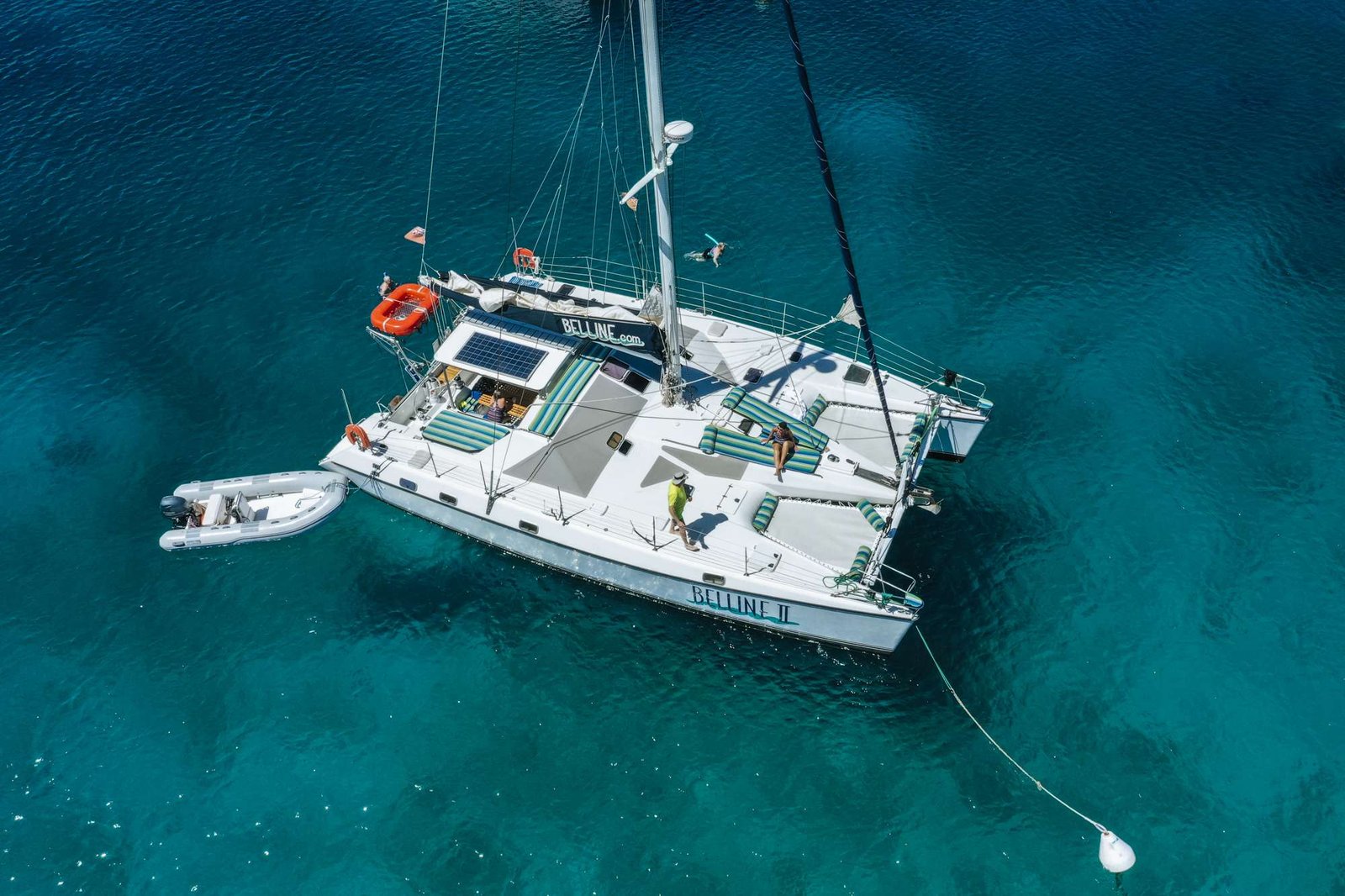 Aerial view of a white catamaran named Belline II in turquoise water, with people sunbathing on deck and a swimmer nearby.