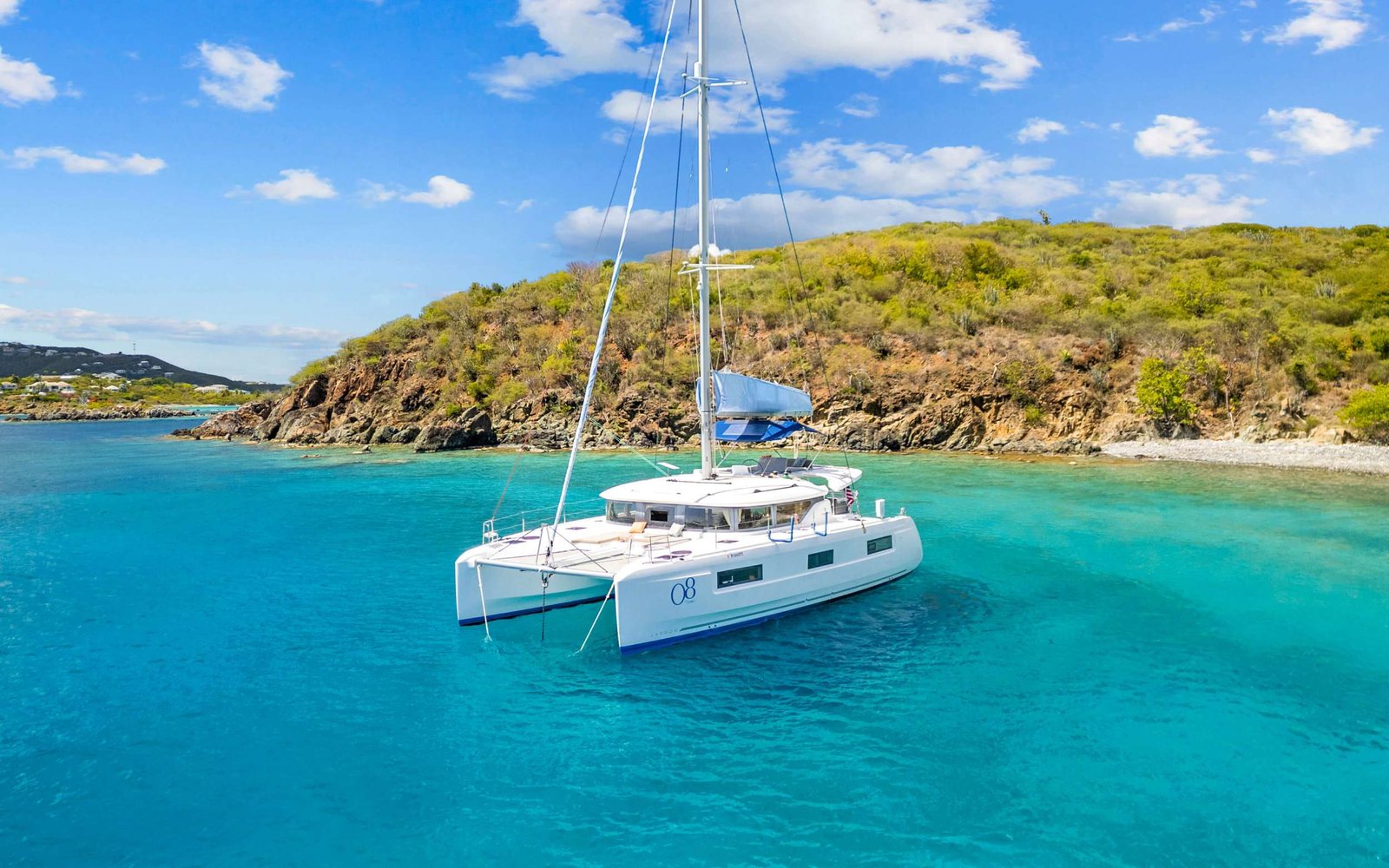 White catamaran moored in turquoise bay with a rocky coastline and green hills under a bright blue sky