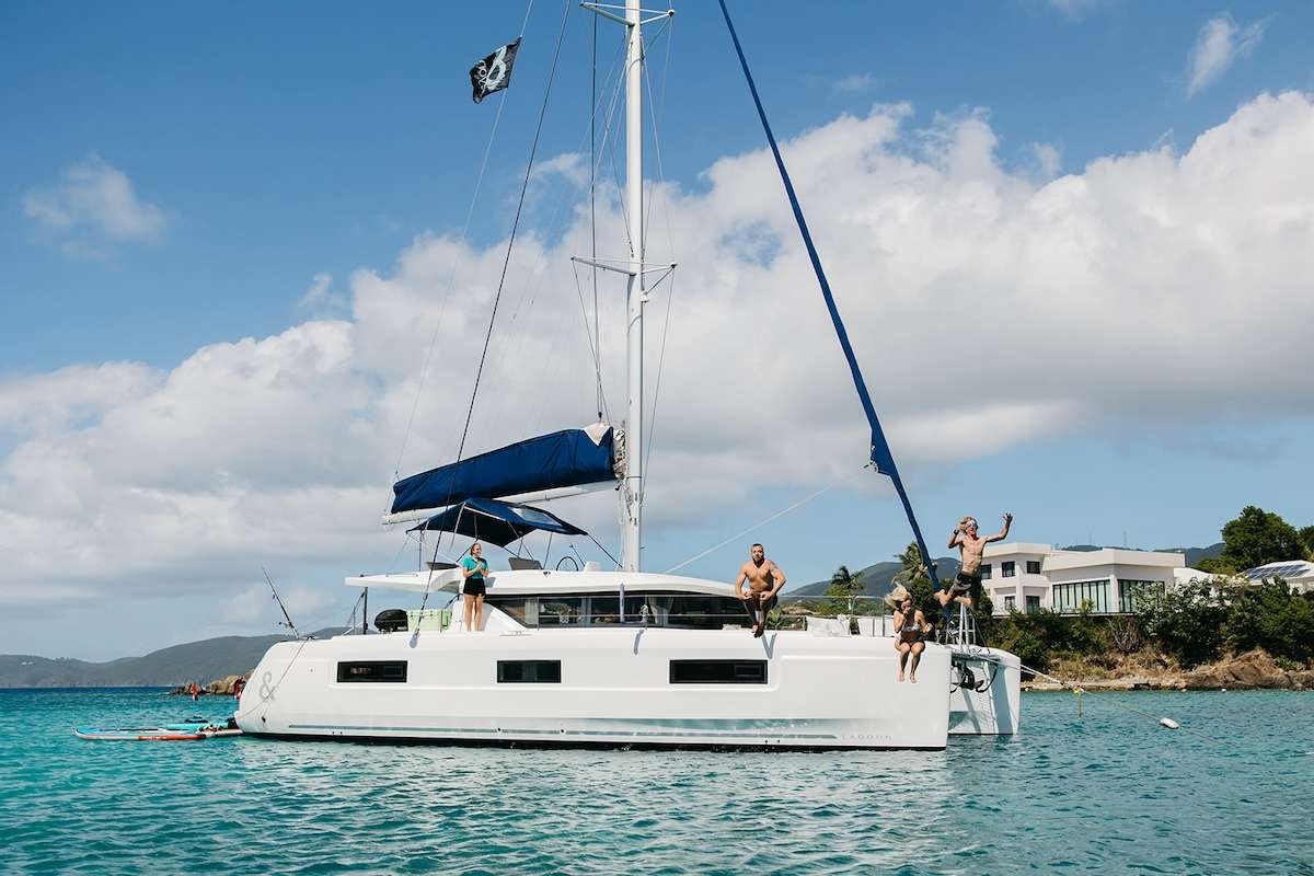 White catamaran anchored on turquoise water with four people on deck; one person jumps into the sea as a bright, sunny day unfolds.