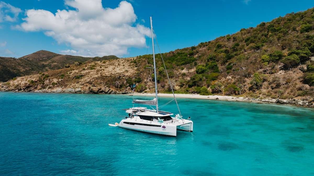 White catamaran anchored in clear turquoise water near a rocky beach with green hills under a bright blue sky.
