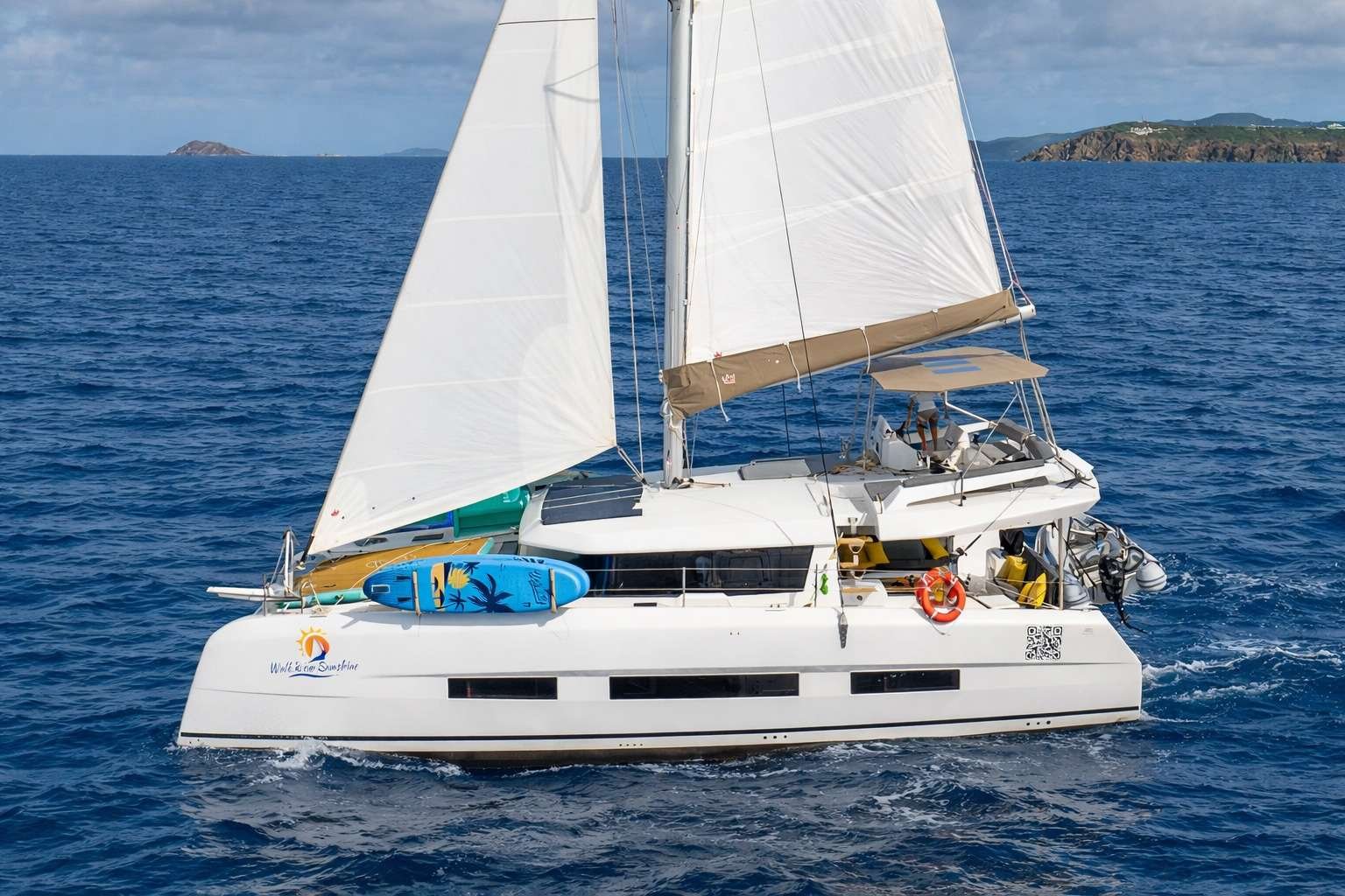 White catamaran sailing boat with two large sails on blue sea; surfboard on bow and crew at cockpit under shade.