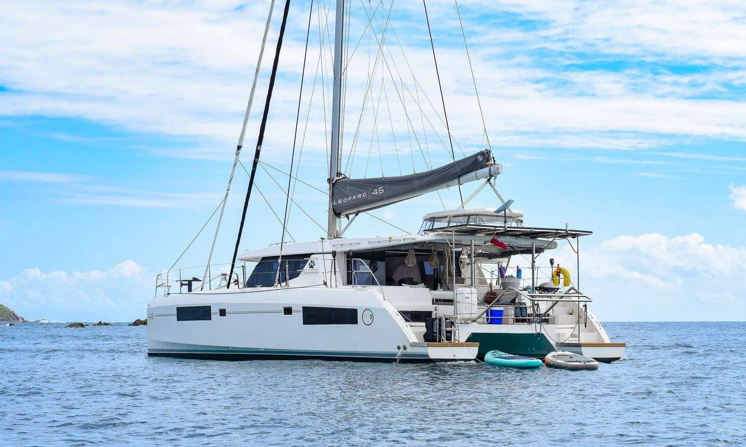 White sailing catamaran anchored on calm blue water under a bright sky, with two paddle boards nearby.