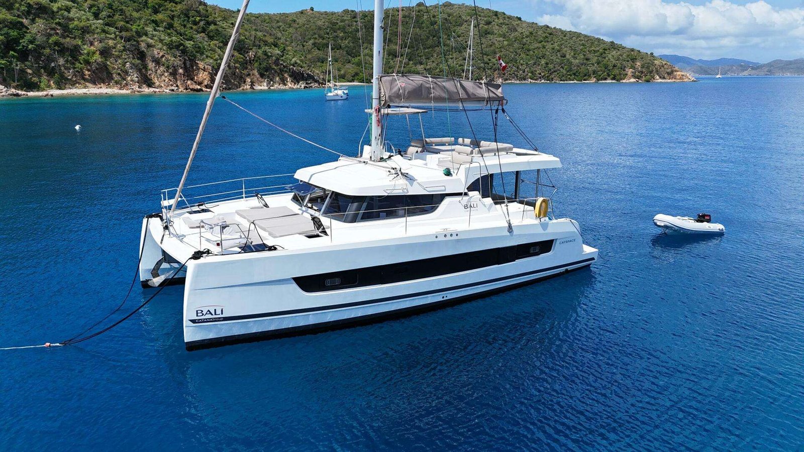 White catamaran anchored in calm blue water near a rocky, green coastline, with a small inflatable dinghy nearby.