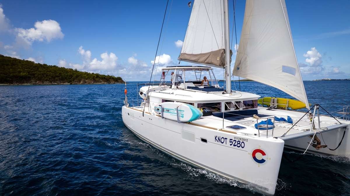 White sailing catamaran on deep blue water with sails raised beside a rocky green island under a clear sky