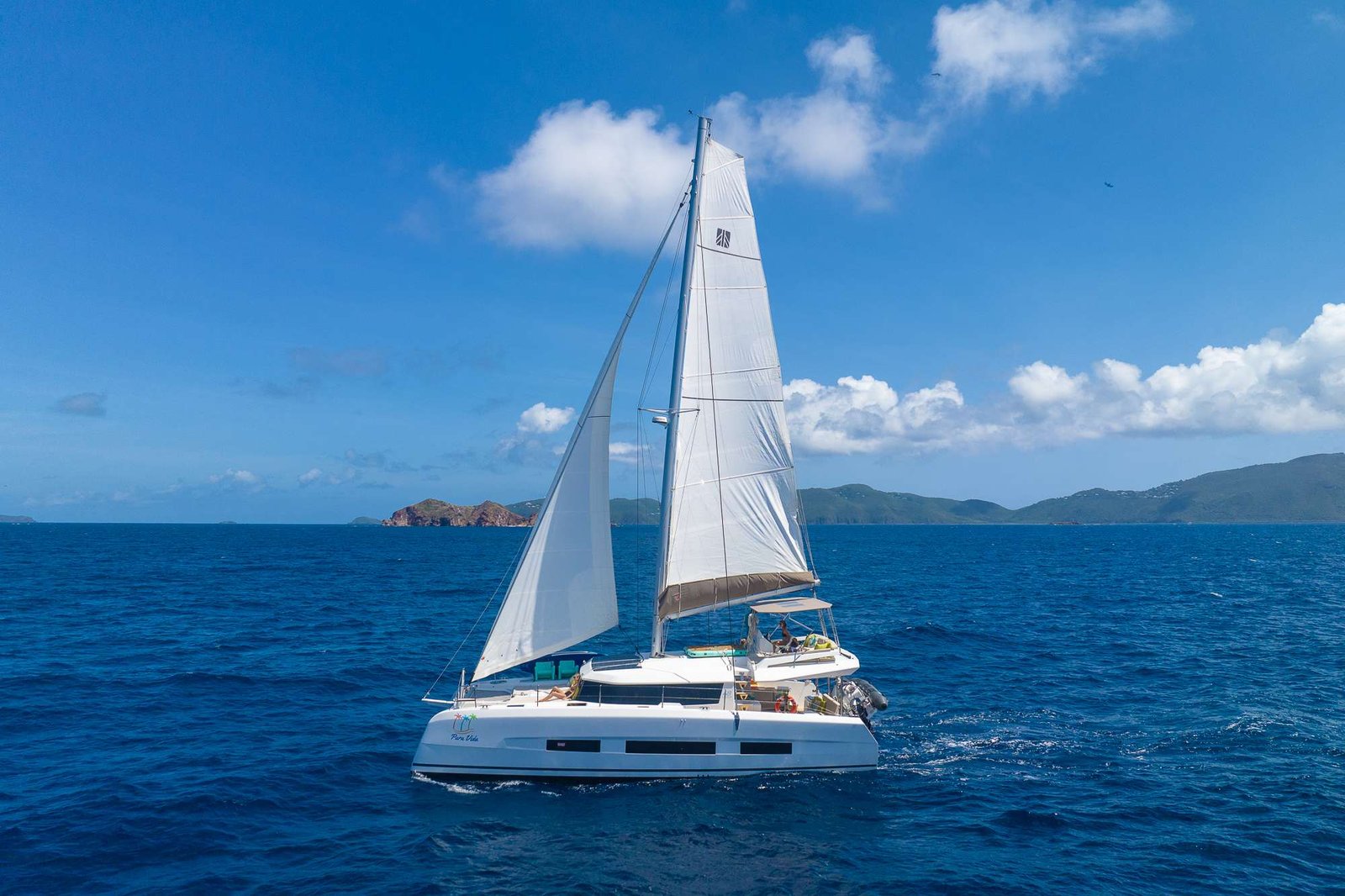 White-sailed catamaran gliding on deep blue ocean with distant green hills on the horizon under a clear sky.