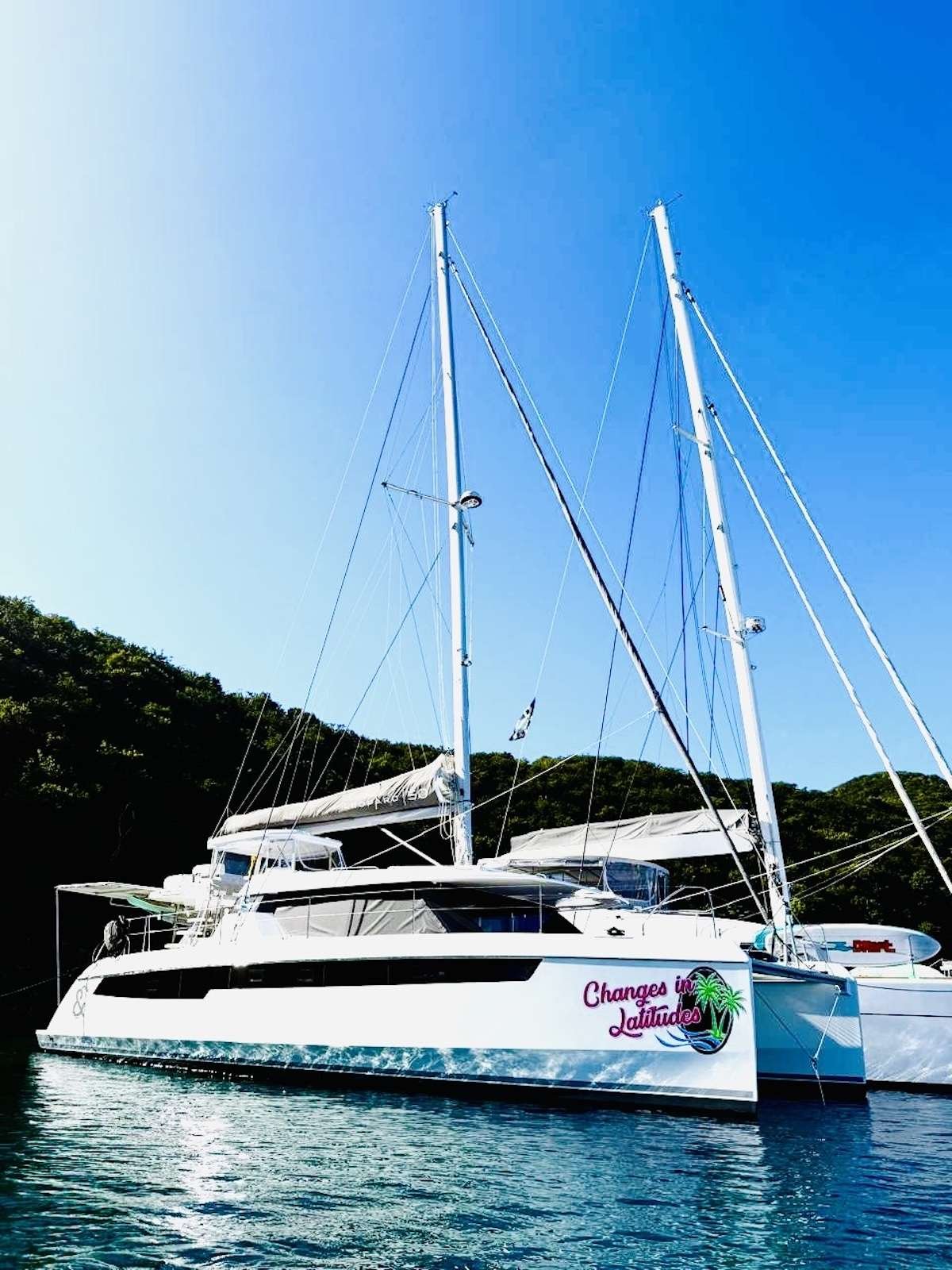 White catamaran yacht docked on calm blue water with two tall masts; hull reads 'Changes in Latitudes' and a palm graphic.