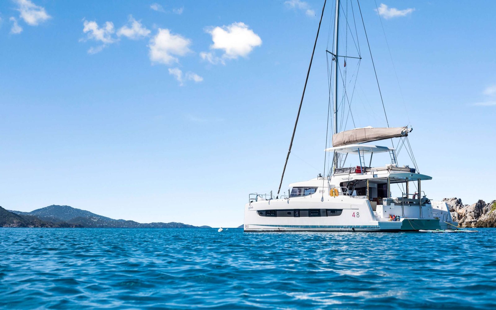 White catamaran moored in calm blue sea with rocky coastline and clear sky in the background.