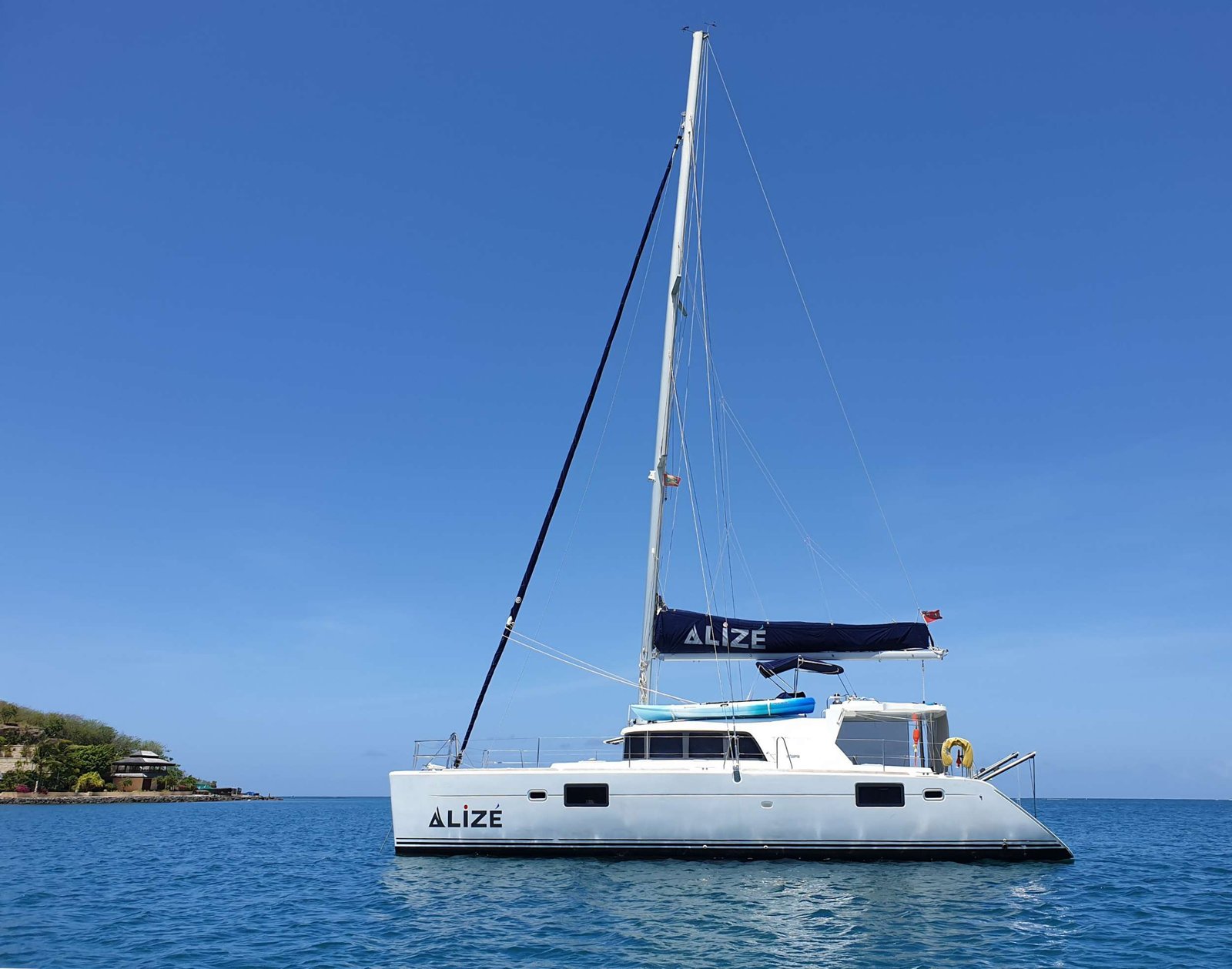 White sailing catamaran named ALIZE on calm blue water with a clear blue sky and coastline to the left