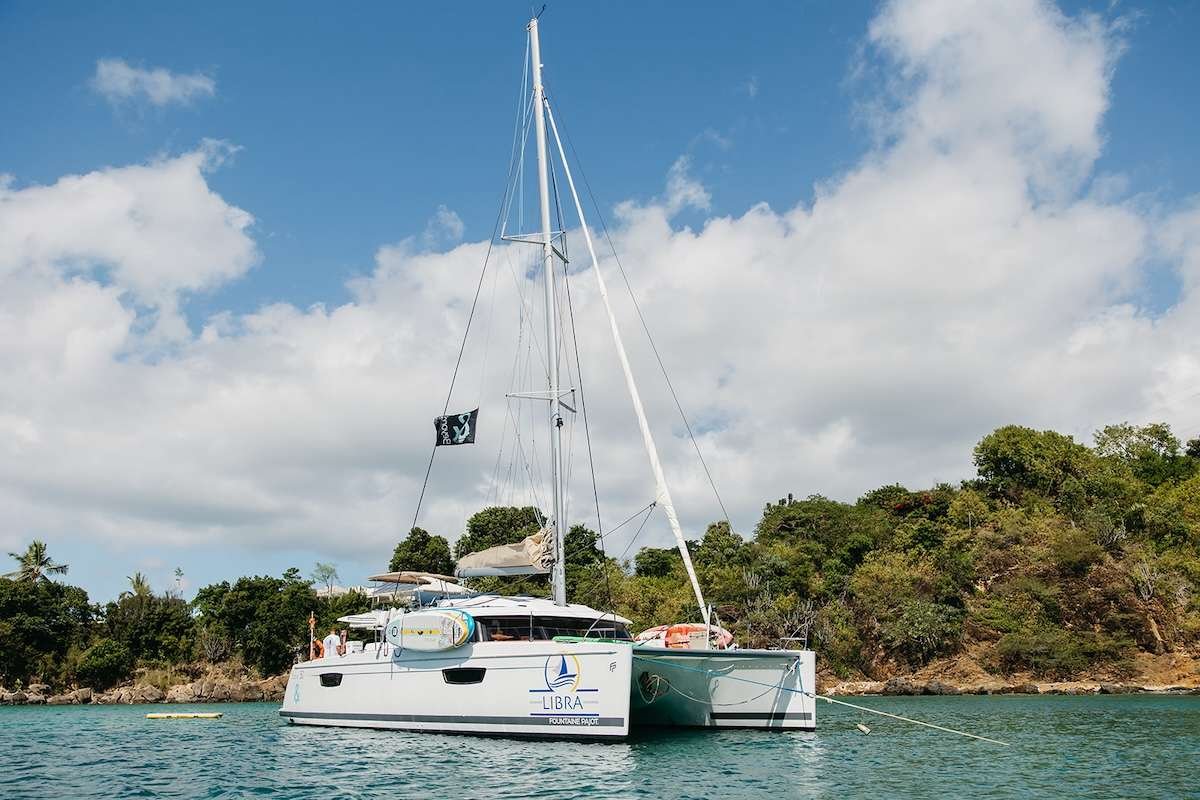 Catamaran Libra anchored in calm turquoise water, with a tall mast and a forested rocky shoreline in the background.