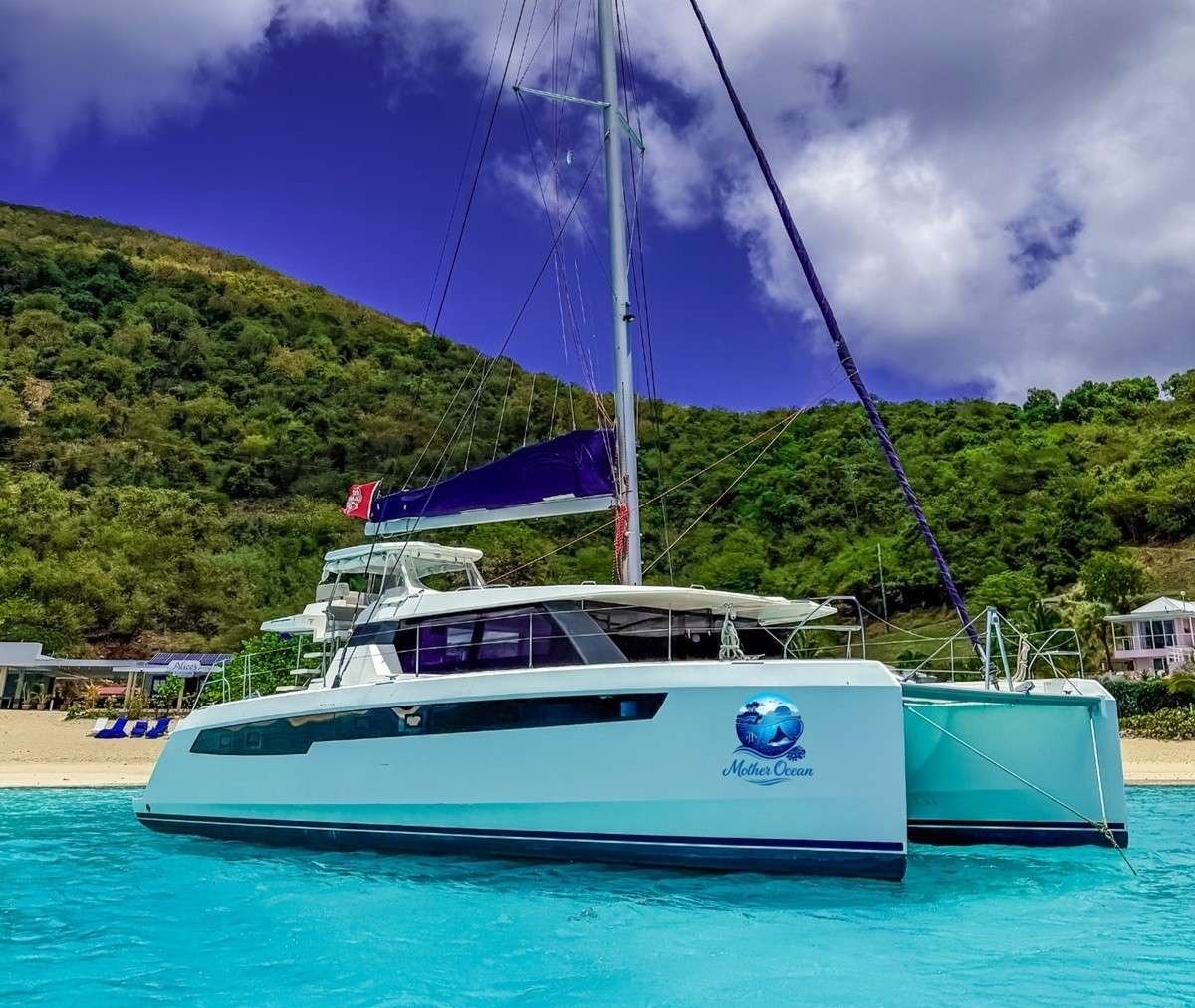 White catamaran named 'Mother Ocean' anchored in turquoise sea with green hills and a sandy beach behind.