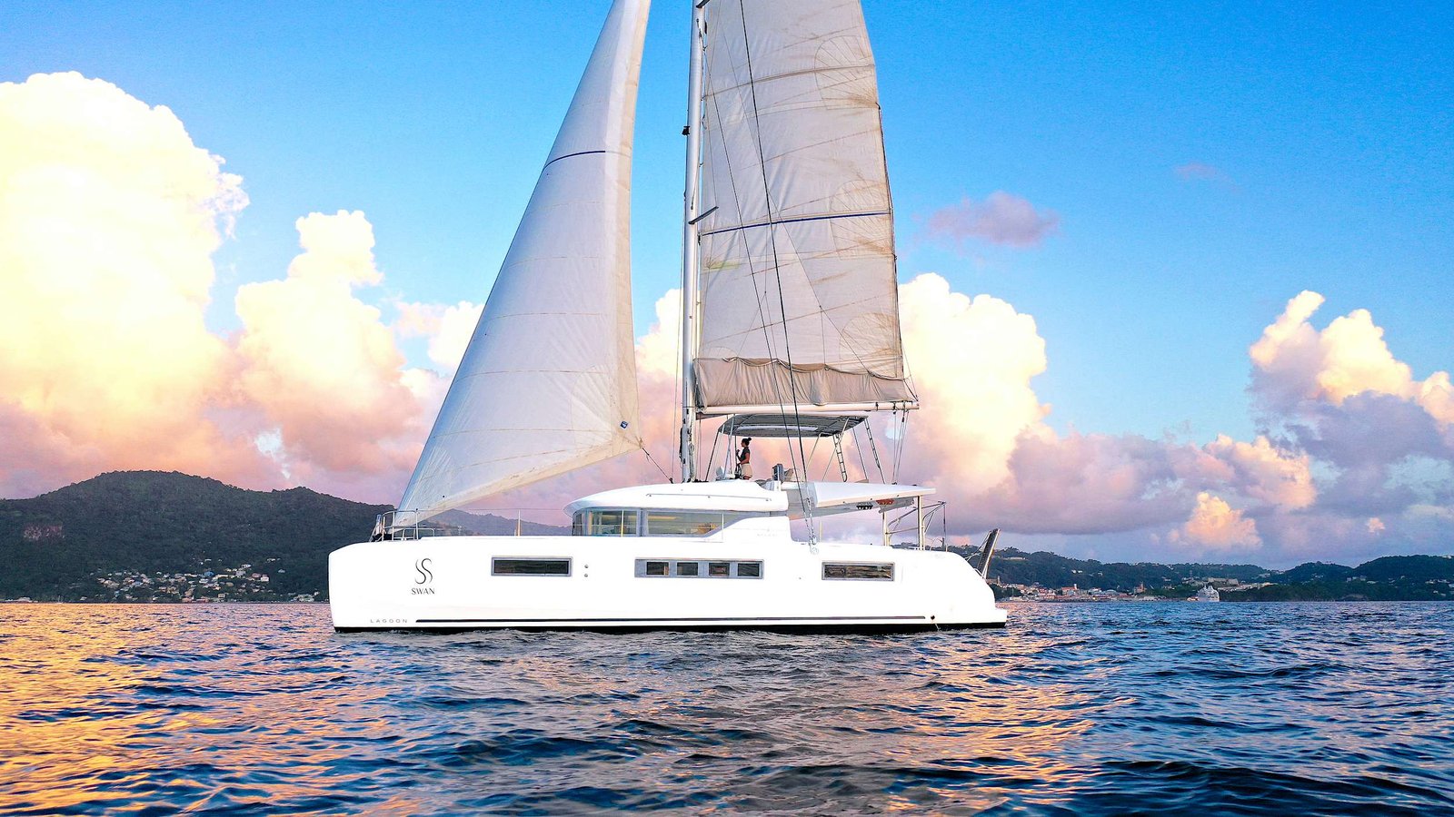 White catamaran sailboat on calm blue water with two tall sails and a coastal town in the distance as sunset clouds glow pink and orange.
