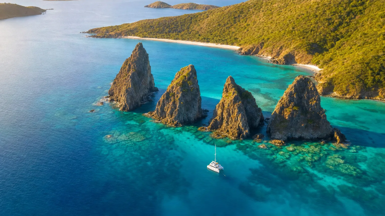 A small white sailboat anchors near jagged sea stacks off a turquoise bay with a forested coastline in the background