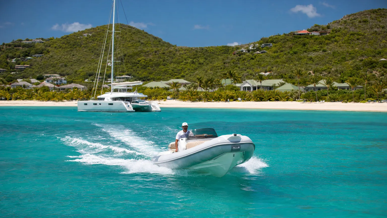 Yacht tender running toward a chic white-sand St. Barts beach with a moored catamaran in a turquoise bay