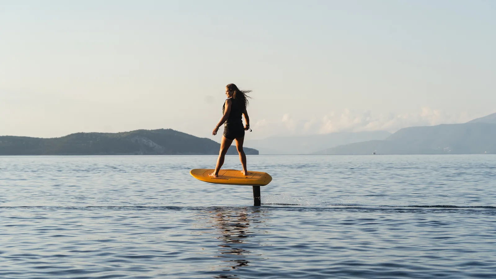 A rider on a yellow Fliteboard eFoil glides above calm water at golden hour