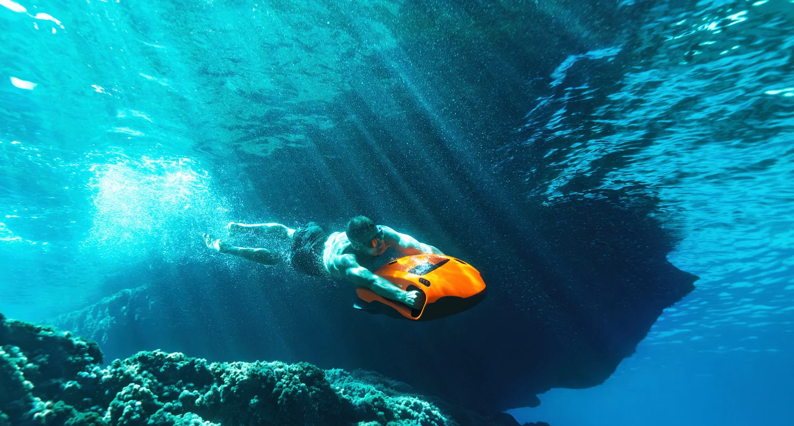 A rider steers an orange SEABOB F9 underwater past a reef and the mouth of a sea cave